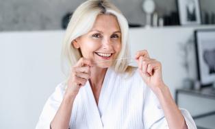 Ältere, lächelnde Frau mit weißem Bademantel benutzt Zahnseide; im Hintergrund ein modernes, helles Badezimmer mit Deko und Bildern