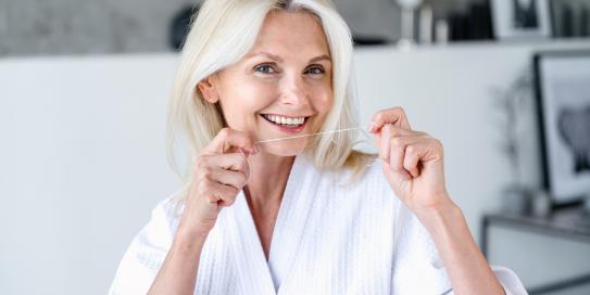 Ältere, lächelnde Frau mit weißem Bademantel benutzt Zahnseide; im Hintergrund ein modernes, helles Badezimmer mit Deko und Bildern