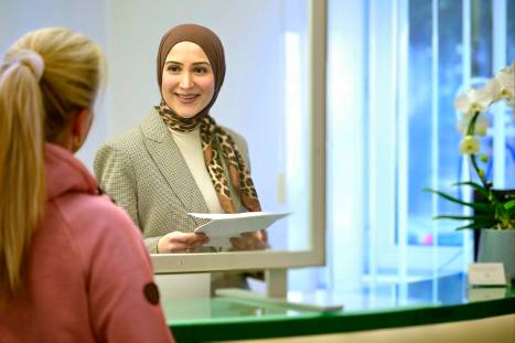 Freundliche Mitarbeiterin mit Kopftuch und Leopardenmuster-Schal steht am Empfangsschalter einer Arztpraxis und überreicht einer Patientin ein Formular