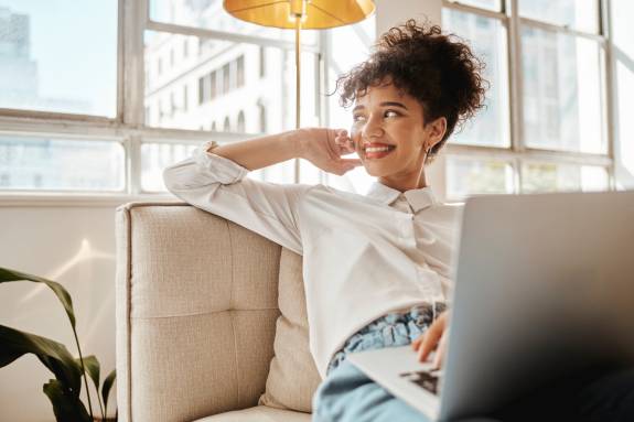 Junge Frau sitzt entspannt auf einem Sofa mit einem Laptop auf dem Schoß, lächelt und blickt aus dem Fenster