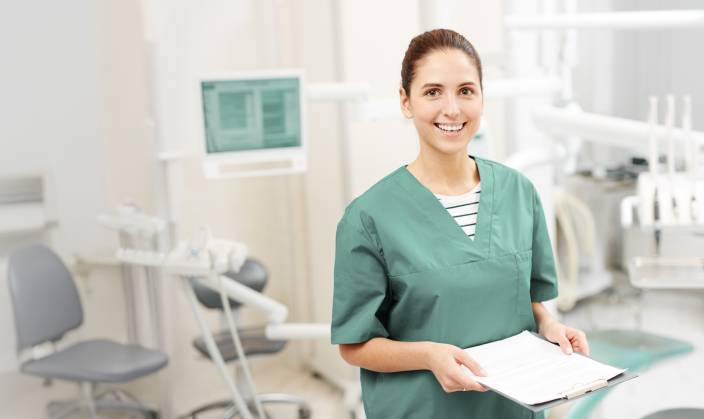 Freundliche Zahnarzthelferin in grüner Kleidung steht mit Klemmbrett in der Hand in einem modernen Behandlungsraum einer Zahnarztpraxis