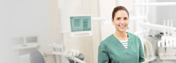 Freundliche Zahnarzthelferin in grüner Kleidung steht mit Klemmbrett in der Hand in einem modernen Behandlungsraum einer Zahnarztpraxis