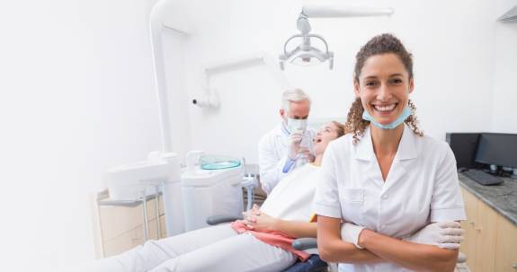 Zahnärztin steht lächelnd mit verschränkten Armen im Vordergrund einer modernen Zahnarztpraxis