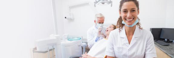 Zahnärztin steht lächelnd mit verschränkten Armen im Vordergrund einer modernen Zahnarztpraxis