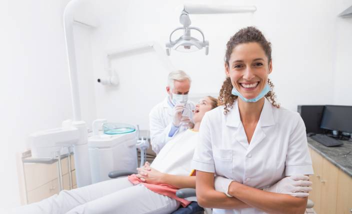 Zahnärztin steht lächelnd mit verschränkten Armen im Vordergrund einer modernen Zahnarztpraxis