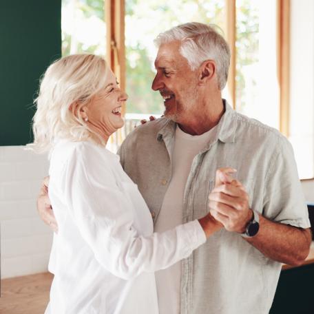Älteres Paar tanzt lachend zusammen in einer hellen, modernen Küche. Beide wirken glücklich und entspannt, während sie sich in die Augen schauen