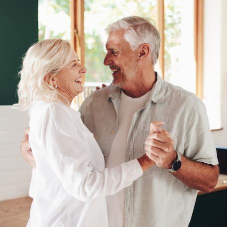 Älteres Paar tanzt lachend zusammen in einer hellen, modernen Küche. Beide wirken glücklich und entspannt, während sie sich in die Augen schauen
