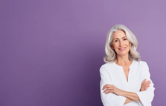 Selbstbewusste ältere Frau mit grauen Haaren und weißer Bluse steht mit verschränkten Armen vor einem lilafarbenen Hintergrund und lächelt freundlich