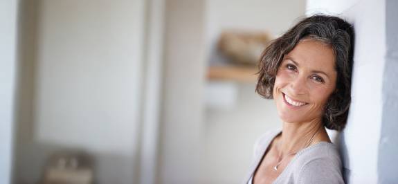 Reife Frau mit schulterlangem, dunklem Haar lehnt lächelnd an einer Wand