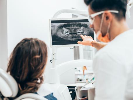 Zahnarzt zeigt einer Patientin ein digitales Röntgenbild ihres Gebisses auf einem Monitor. Beide befinden sich im Behandlungszimmer einer modernen Zahnarztpraxis