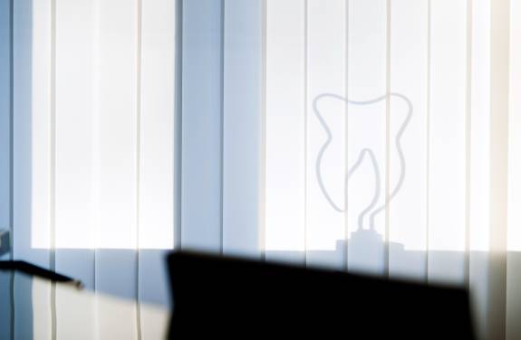 Licht fällt durch vertikale Lamellenjalousien und wirft den Schatten einer stilisierten Zahn-Skulptur an die Wand