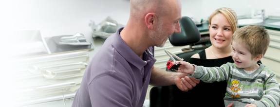 In einer Zahnarztpraxis überreicht ein lachender Junge dem behandelnden Zahnarzt, einem glatzköpfigen Mann in lila Poloshirt, ein Spielzeugauto