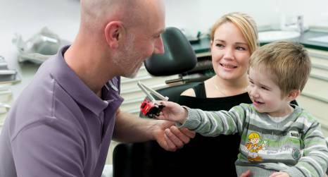 In einer Zahnarztpraxis überreicht ein lachender Junge dem behandelnden Zahnarzt, einem glatzköpfigen Mann in lila Poloshirt, ein Spielzeugauto