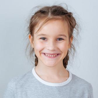 Fröhliches junges Mädchen mit zwei Zöpfen lächelt in die Kamera. Sie trägt ein graues T-Shirt mit weißem Kragen